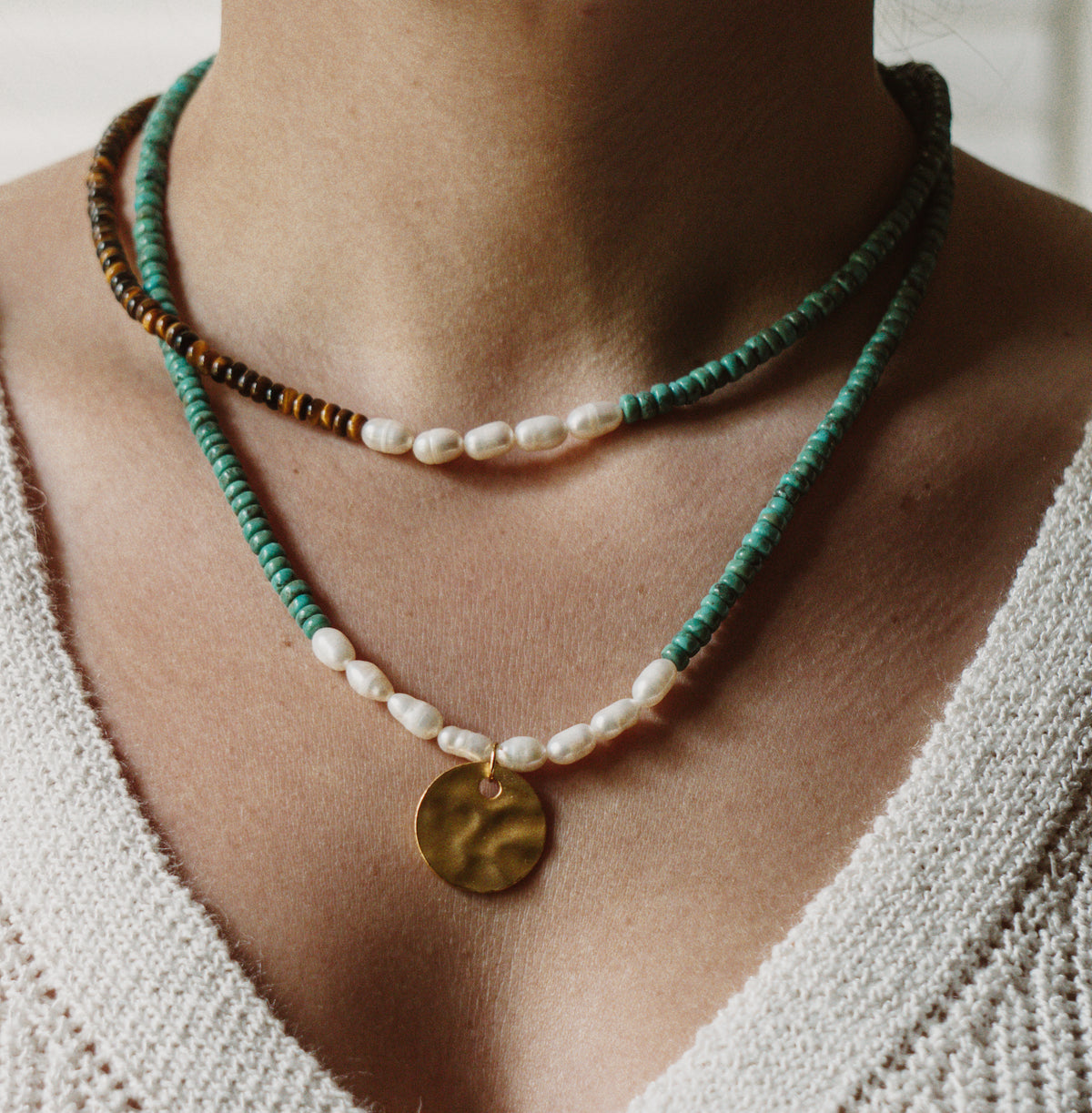 A close-up of a person's neck and chest wearing a layered necklace set. The top layer is a beaded turquoise and brown cord necklace with white pearl-like beads.
