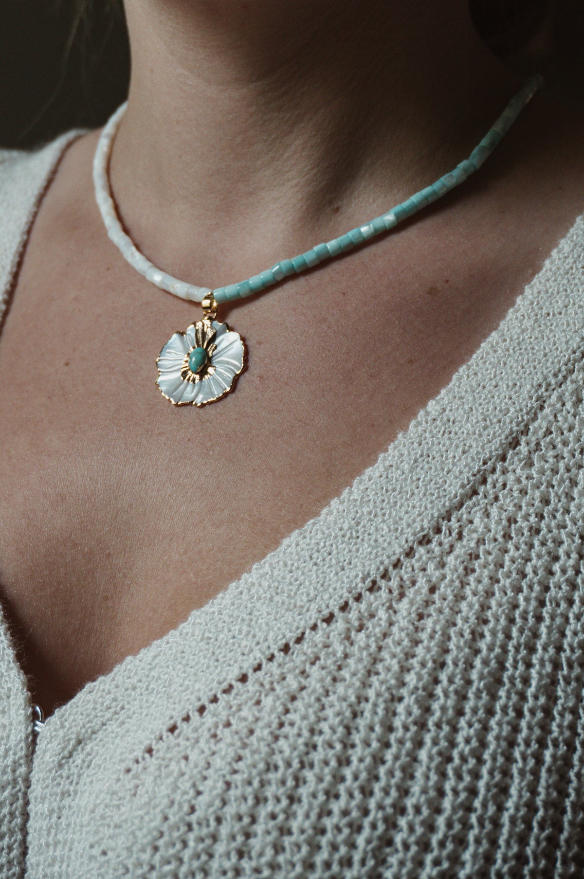 A close-up of a person wearing a delicate necklace with a white flower pendant featuring turquoise and gold details, paired with a textured light gray knit top.